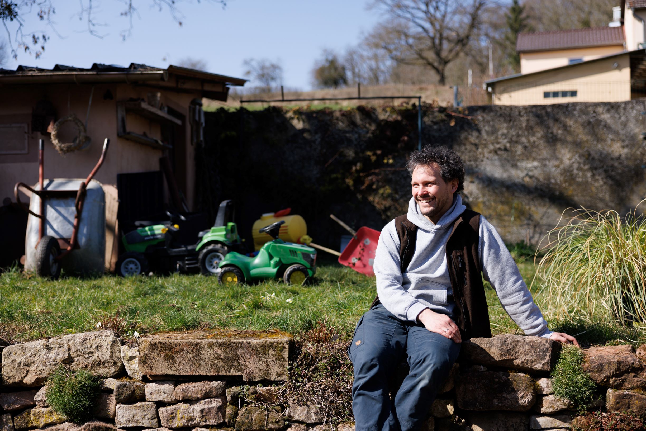 Homme assis sur un muret de jardin, on voit son jardin en arrière-plan avec des outils de jardinage et des jouets d'enfants.