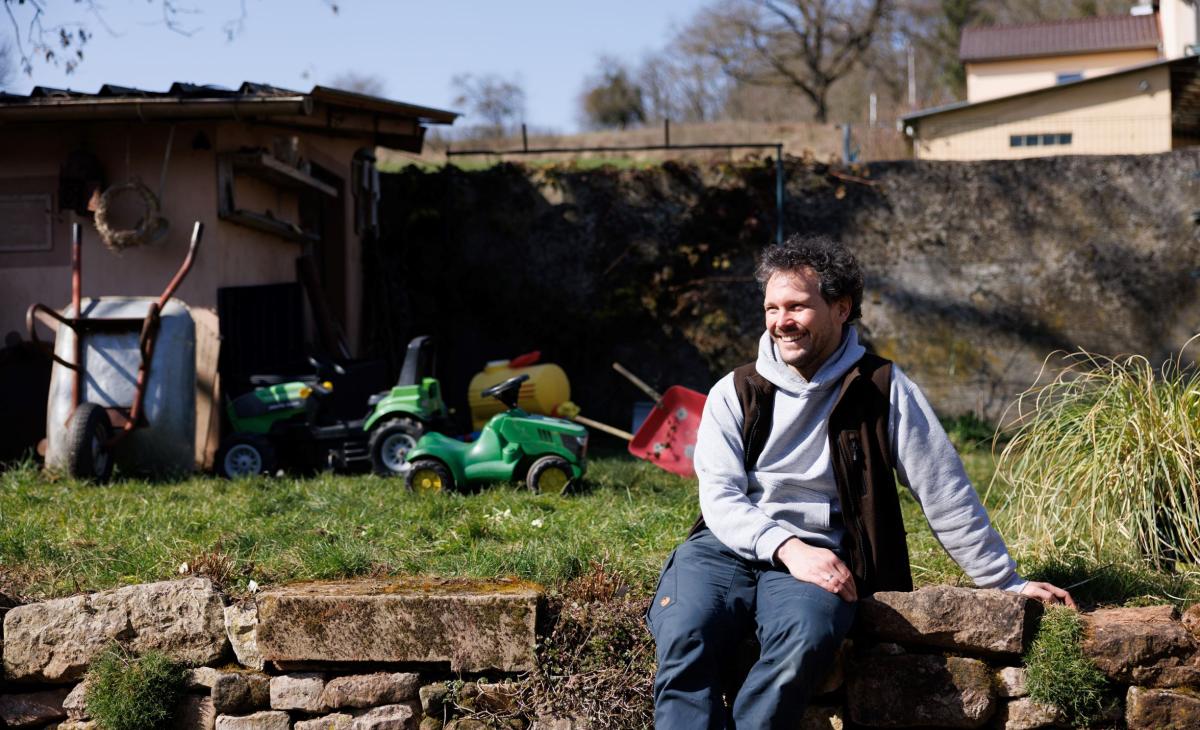 Homme assis sur un muret de jardin, on voit son jardin en arrière-plan avec des outils de jardinage et des jouets d'enfants.